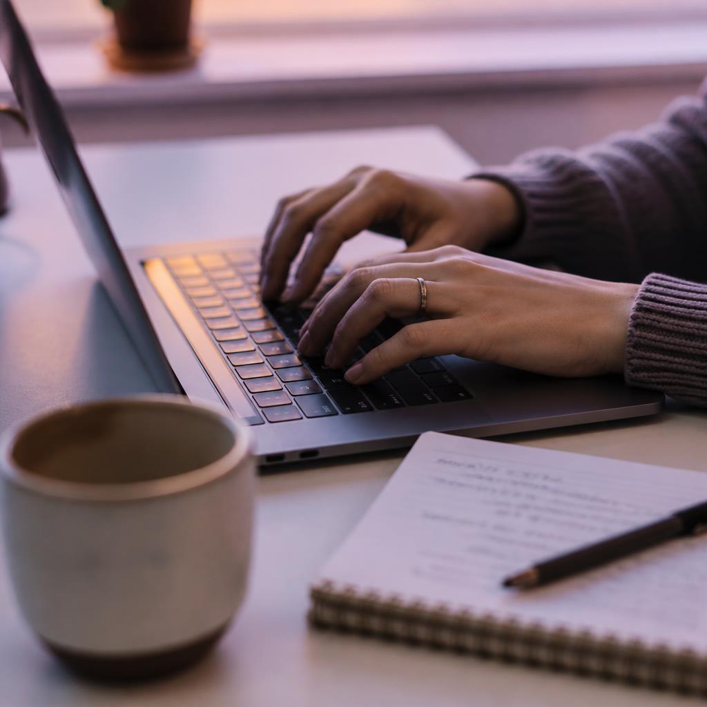 Hände tippen konzentriert auf einem Laptop neben Notizbuch und Kaffee